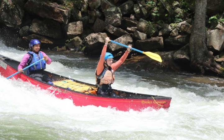 Two people wearing safety gear paddle a canoe through whitewater. The person in the front of the canoe raises their paddle into the air in apparent celebration. 